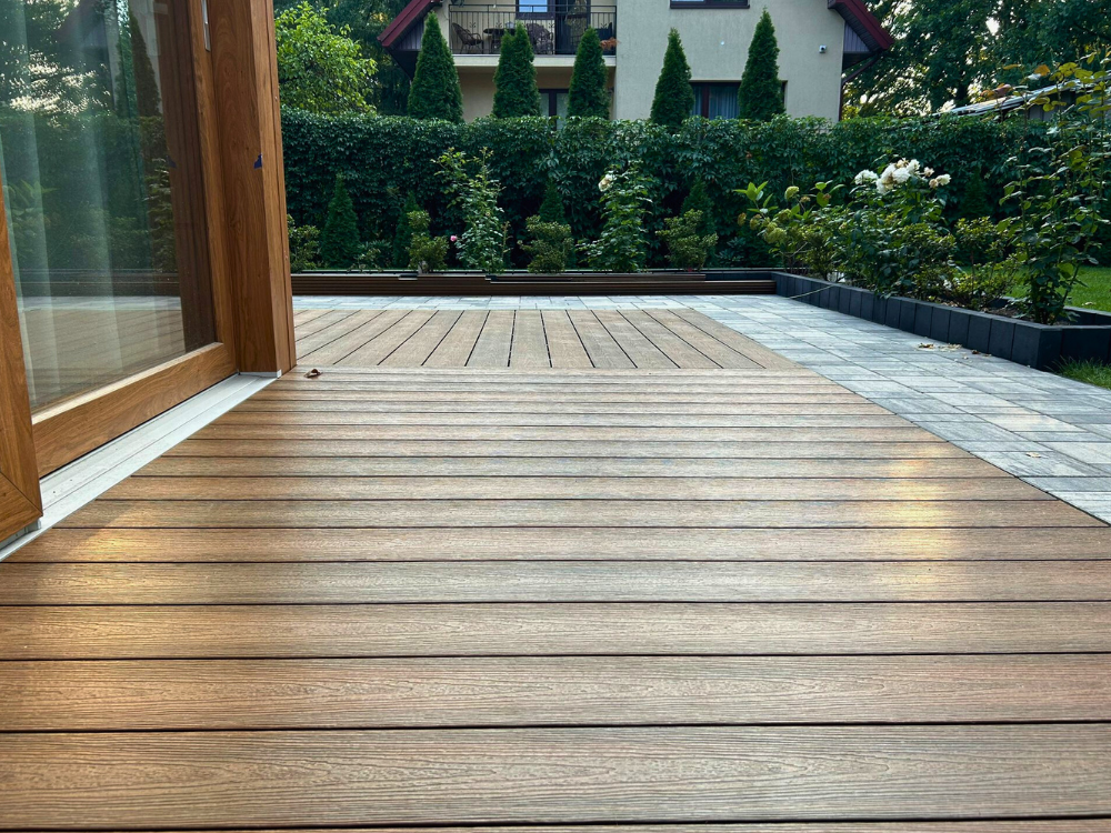 Taras z desek kompozytowych o powłoce polimerowej w kolorze teak, otoczony zielenią w ogrodzie przy nowoczesnym domu.