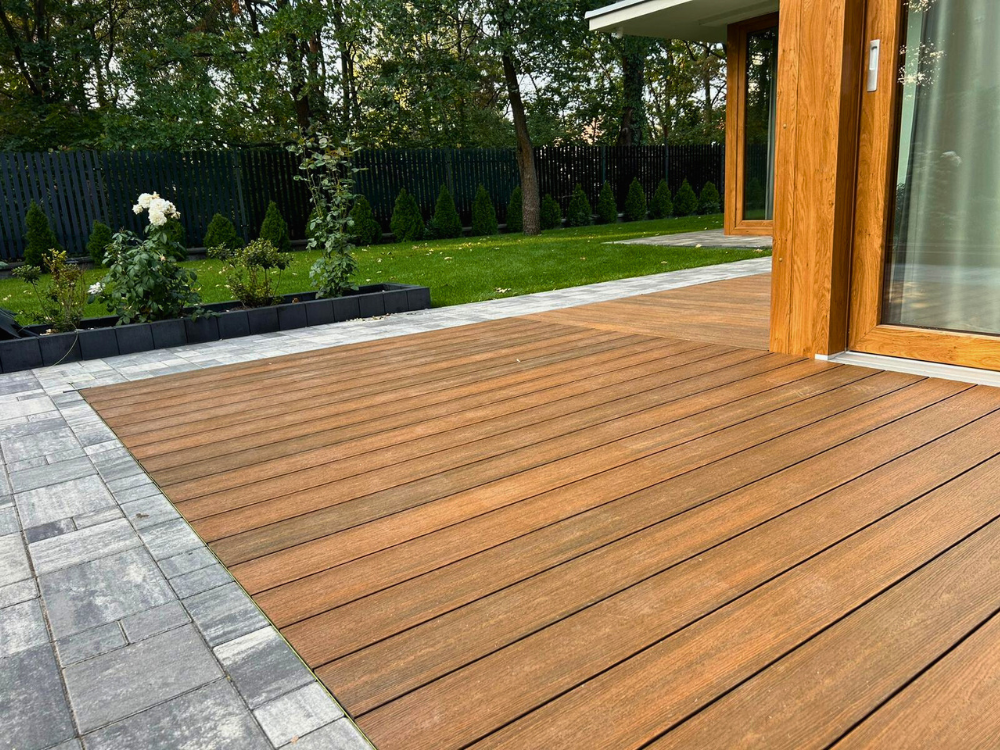 taras kompozytowy z desek Advance (2) Nowoczesny taras z desek kompozytowych w kolorze drewna, otoczony ogrodem z zielenią i kostką brukową